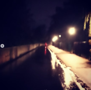 Reflet de nuit d'une personne avec un manteau rouge sur un vélo, sur une piste cyclable mouillée par la pluie et éclairée par des lampadaires.